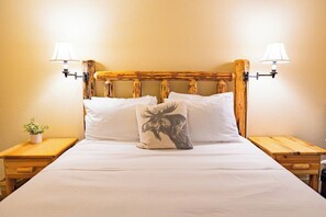 Ein Bett mit weißer Bettwäsche, einem Holz-Kopfbrett, zwei Nachttischlampen und einem Zierkissen mit Elch-Motiv.