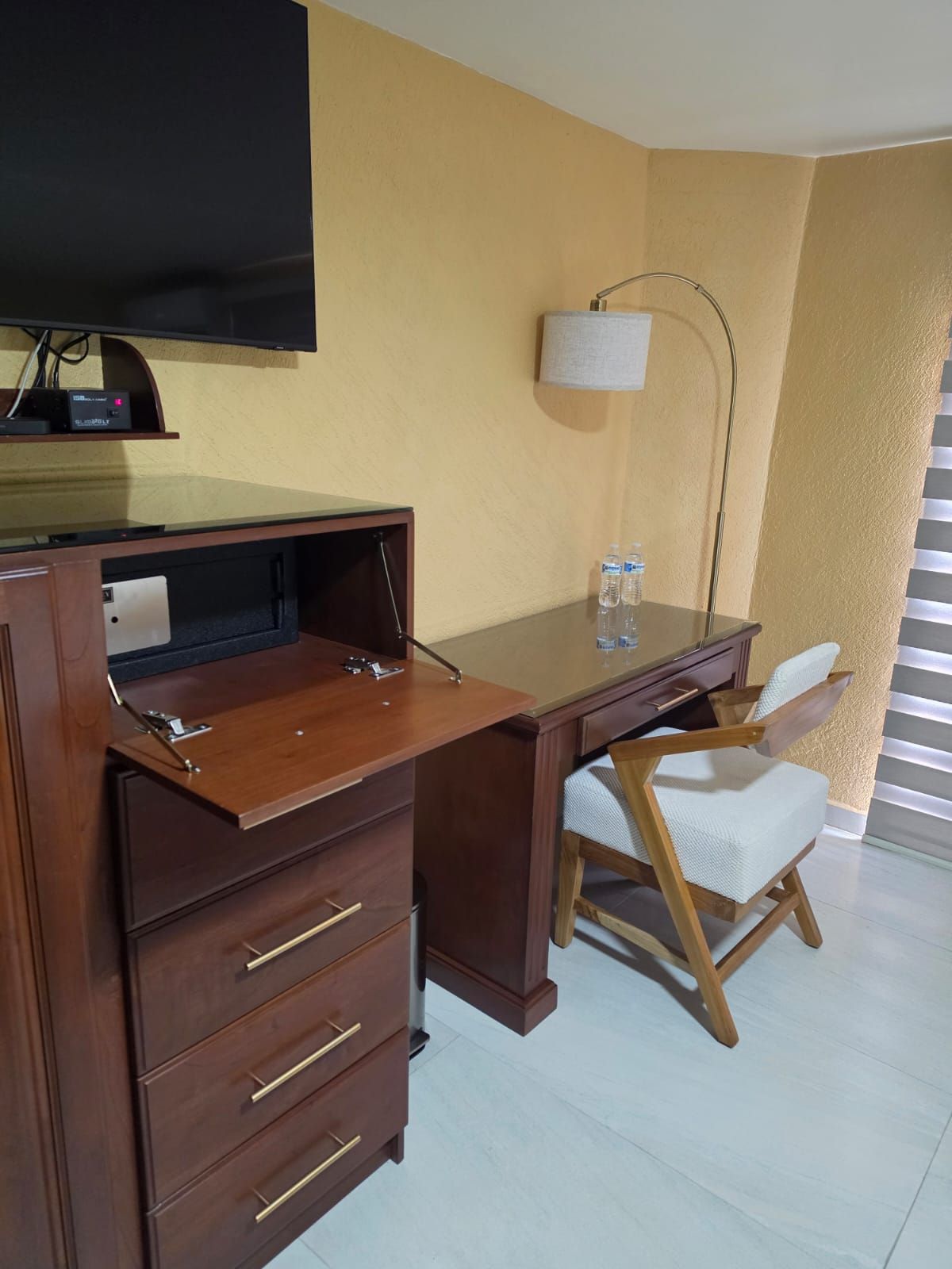 A hotel room with a desk, chair, television, lamp, and a water bottle.