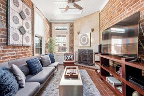 A living room with a brick wall, a sofa, a coffee table, a fireplace, and a television. - The Lyons Estate | Historic Savannah Luxury (Savannah)