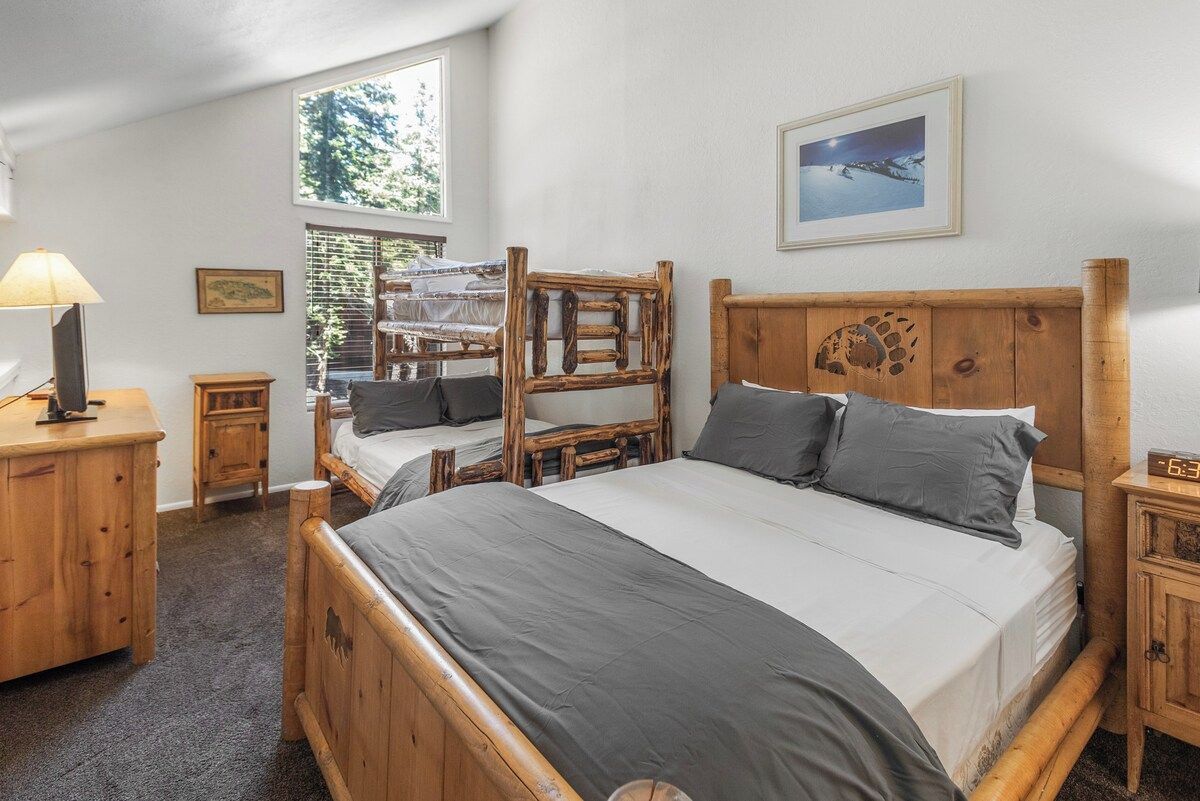 A room with a large bed, two smaller beds, a wooden desk, a nightstand, and a window with a view of trees.