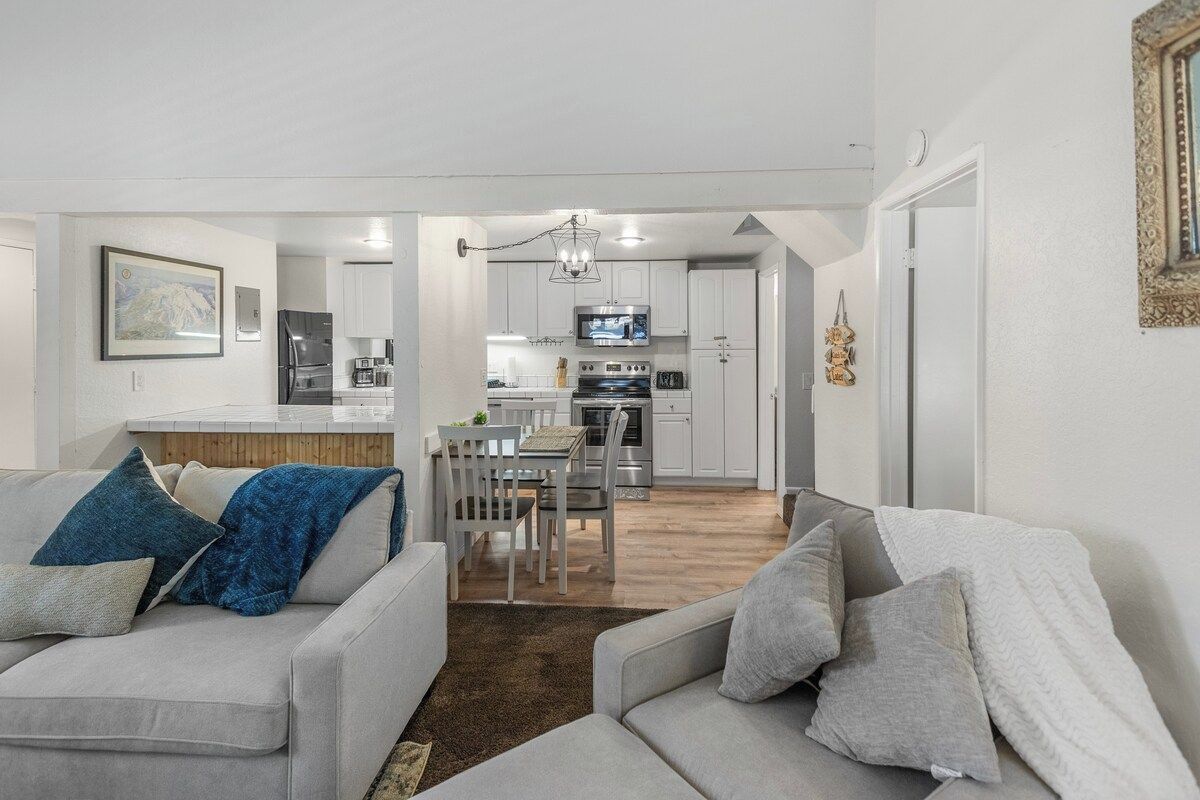 A modern living room with a sofa and a dining table, a kitchen with white cabinets and appliances, and a hallway leading to another room.