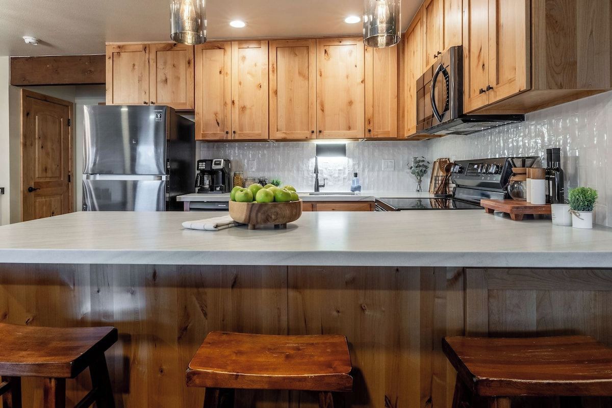 A kitchen with wooden cabinets, a stainless steel refrigerator, a stove, and a large island with a bowl of green apples.