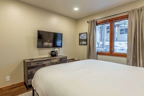 Ein Schlafzimmer mit einem Bett, einem an der Wand befestigten Fernseher, einer Kommode und einem Fenster mit Vorhängen.