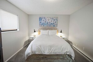 Een slaapkamer met een bed, twee nachtkastjes, een raam met jaloezieën en een wanddecoratie.
