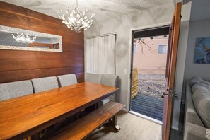 Een eetgedeelte met een houten tafel en banken, een kroonluchter en uitzicht door een deur op een buitenruimte.