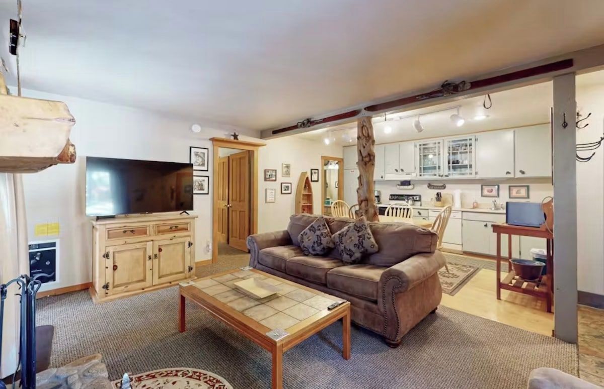 A living room with a sofa, coffee table, and a television. There is a kitchen area with cabinets and a dining table.