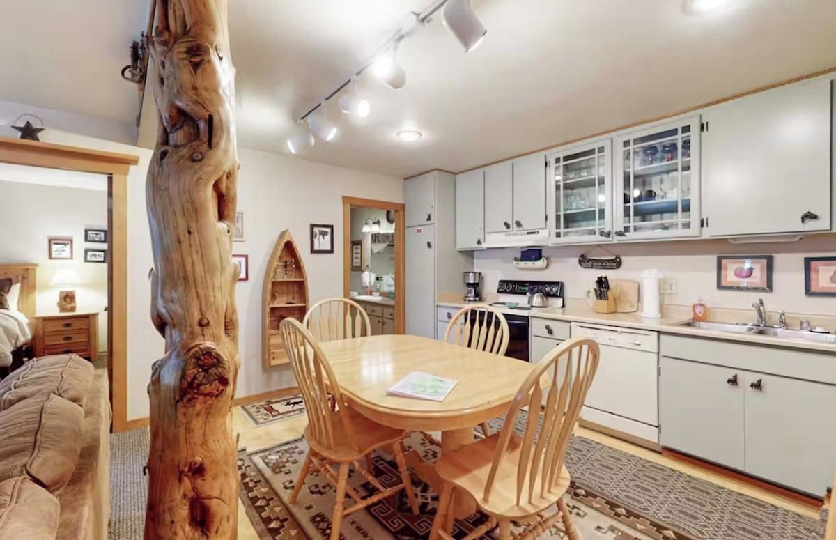 Un coin repas avec une table et des chaises en bois, une cuisine avec des meubles blancs et un tronc d’arbre visible.