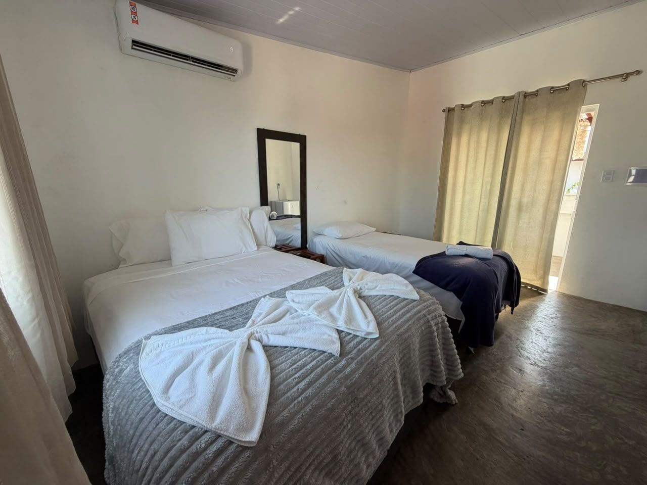 A hotel room with two beds, a mirror, a window with curtains, and a suitcase.