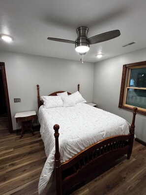 A bedroom with a wooden bed, a nightstand, a ceiling fan, and a window with a picture frame. - Welcome to Pymatuning Lake Lodge - a 3 Bedroom, 2 Bathroom getaway! (Pymatuning Central)