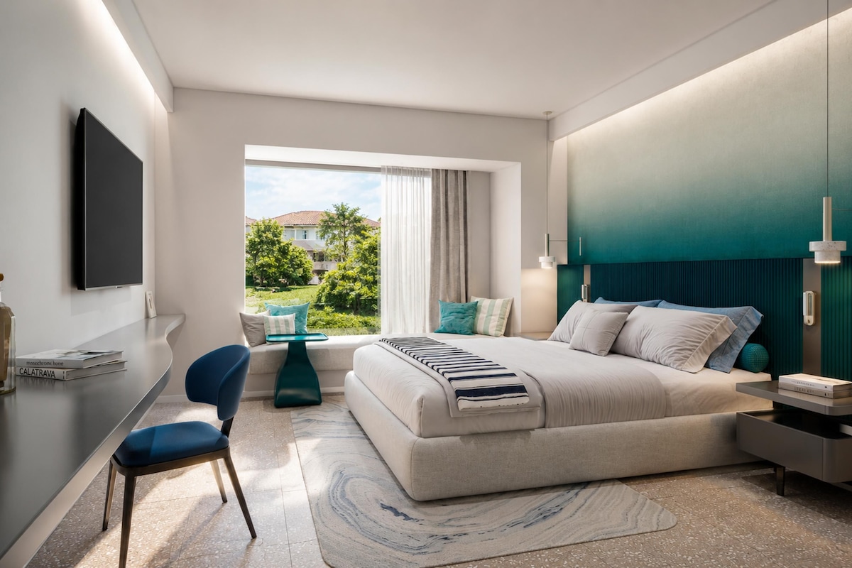 Ein modernes Schlafzimmer mit einem großen Bett, einem Schreibtisch und einem Stuhl. Ein Fenster bietet Blick auf grüne Natur.