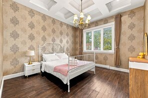 A bedroom with a white metal bed, a nightstand with a lamp, a window with curtains, and a chandelier. - Tranquil 2-bedroom house in charming Surrey perfect for relaxing stays (Surrey)