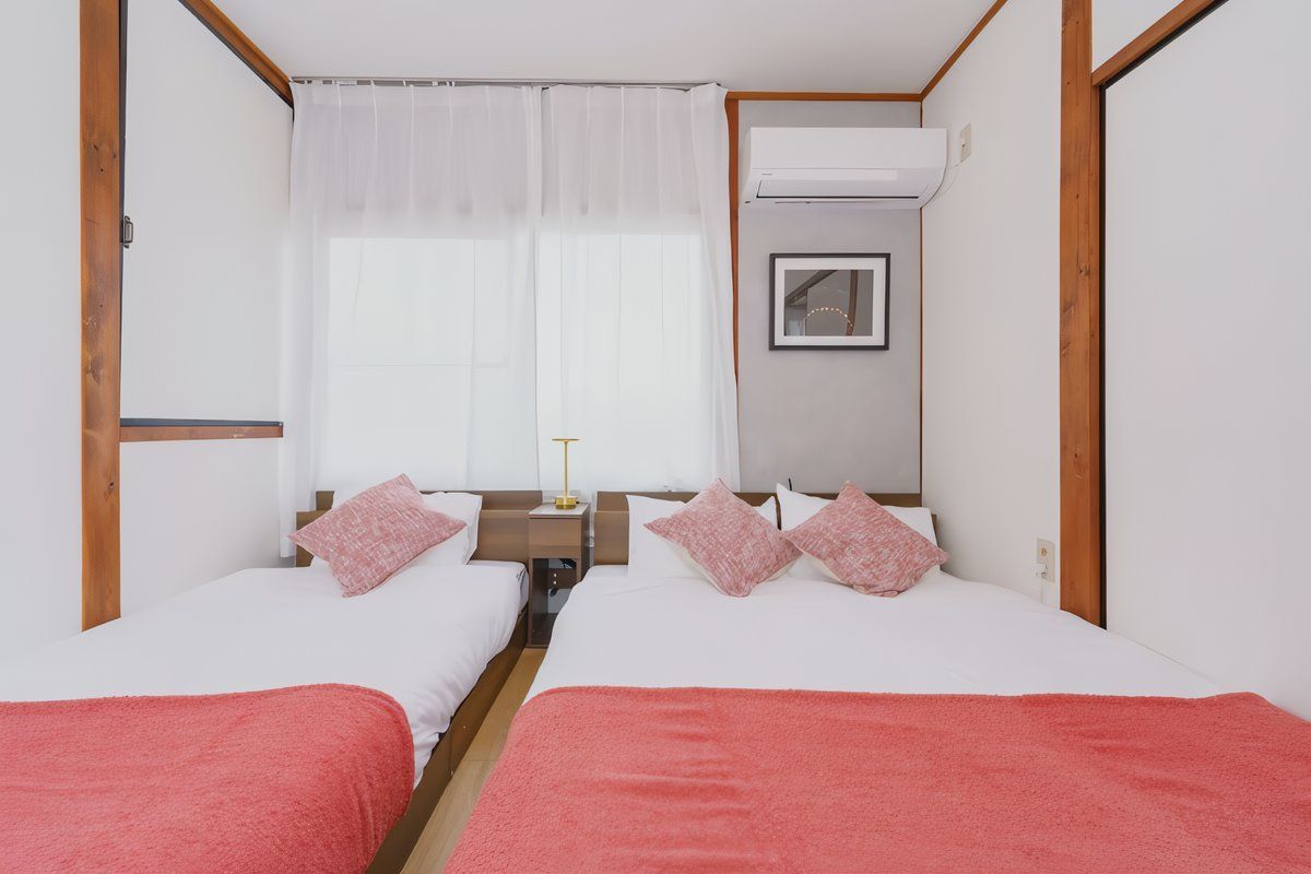 A double room with two beds, a bedside table with a lamp, a window with curtains, and an air conditioning unit.