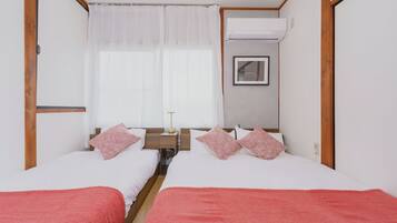 A double room with two beds, a bedside table with a lamp, a window with curtains, and an air conditioning unit.