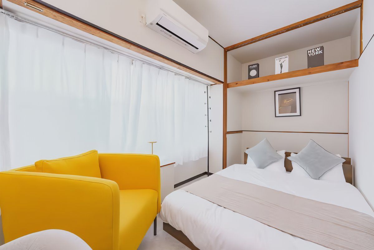 A hotel room with a bed, a yellow armchair, a bedside table, a wall-mounted air conditioner, and a shelf with framed pictures and a clock.