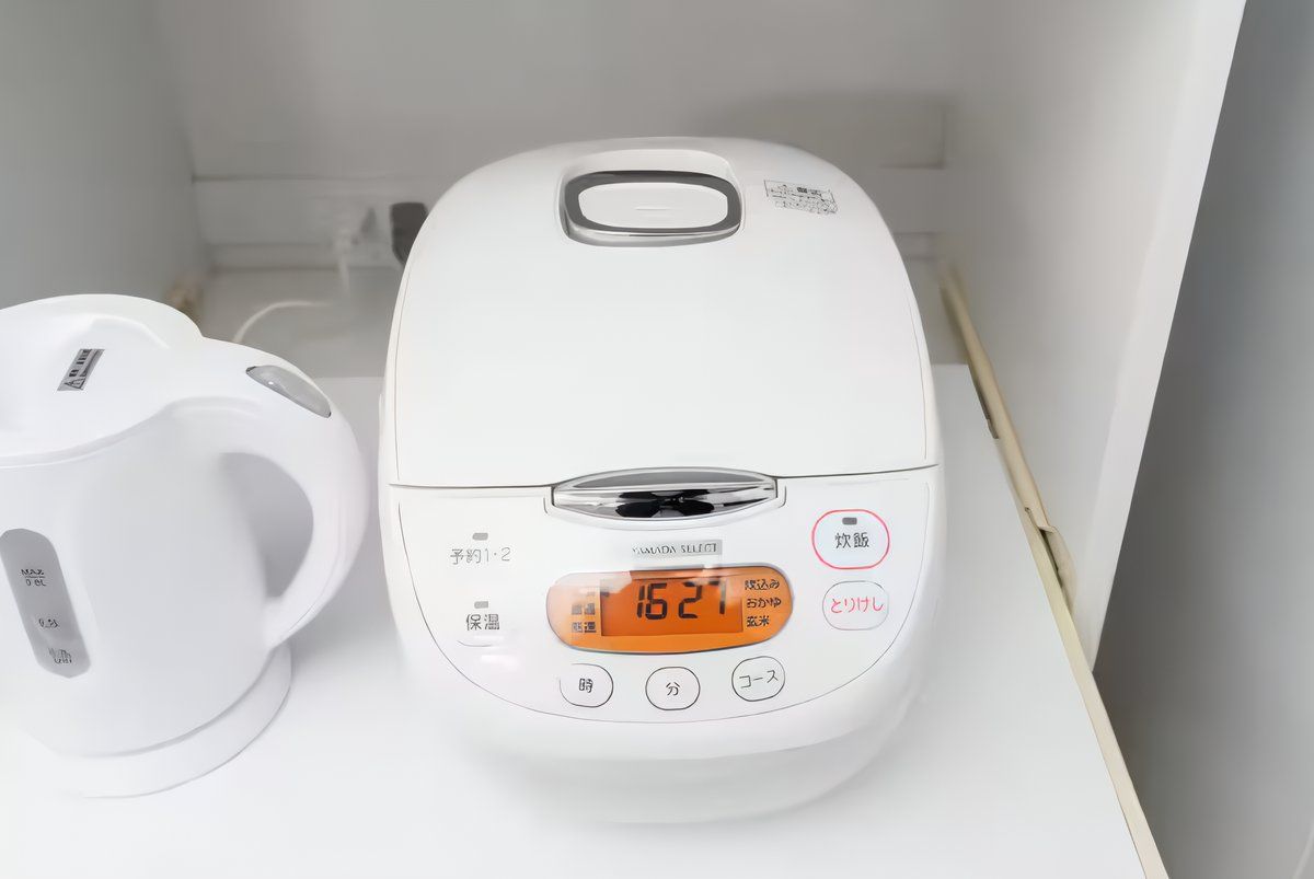 A rice cooker with a digital display and buttons, placed next to a white kettle on a white surface.
