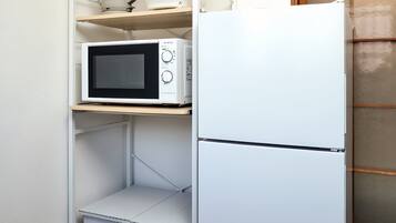 A compact kitchen with a white refrigerator, a microwave, and a kettle on a shelf.