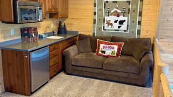 A compact kitchen with wooden cabinets, a microwave, and a sink. A brown sofa with a 'Big Bear Campground' pillow is in the living area.
