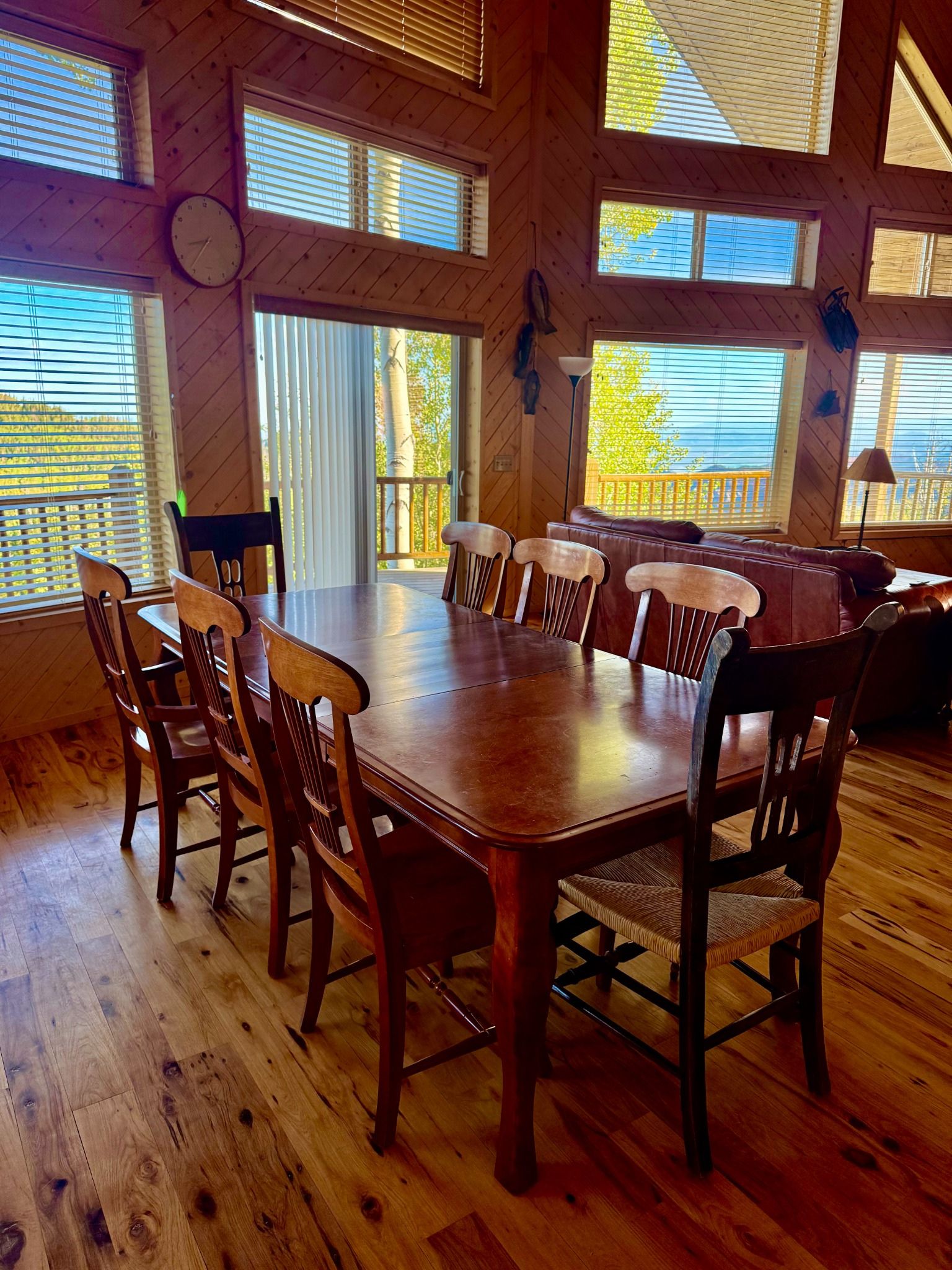 Un comedor de madera con una mesa grande y sillas, ventanas amplias con persianas y vistas al exterior.