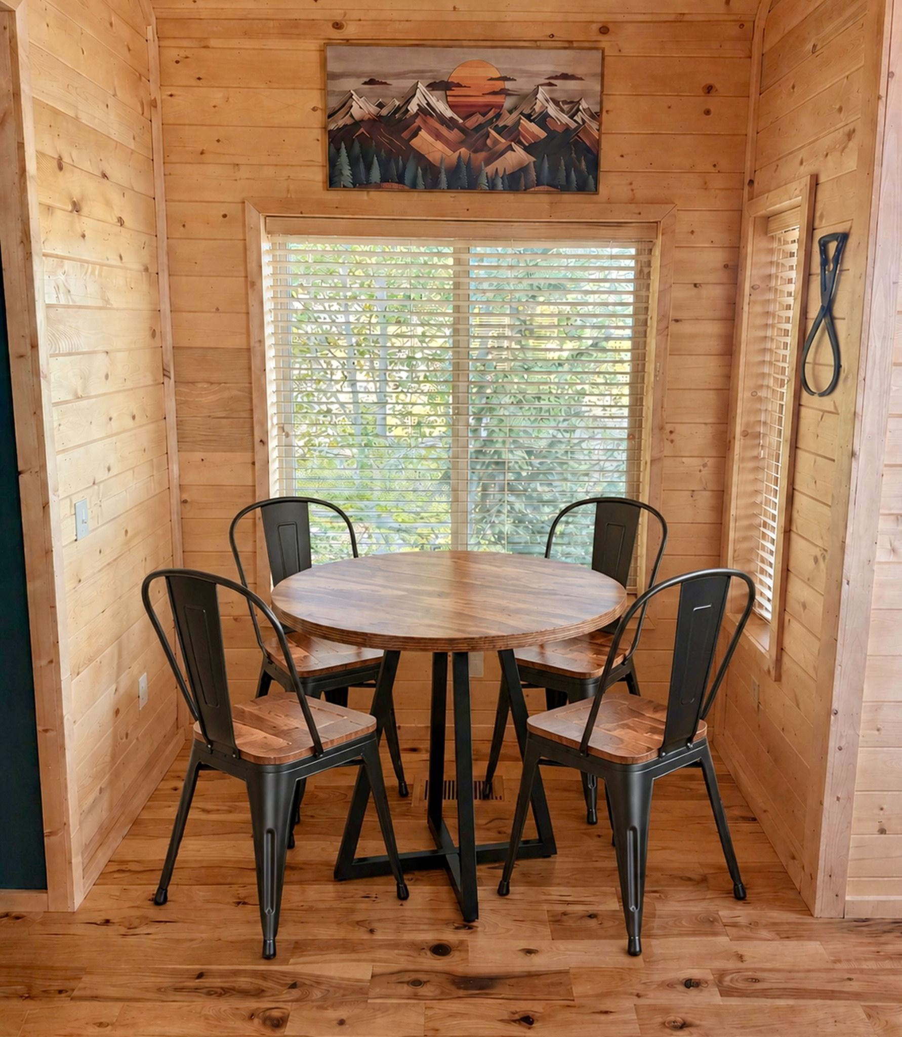 A wooden interior with a round table and four chairs, a large window with blinds, and a painting on the wall.