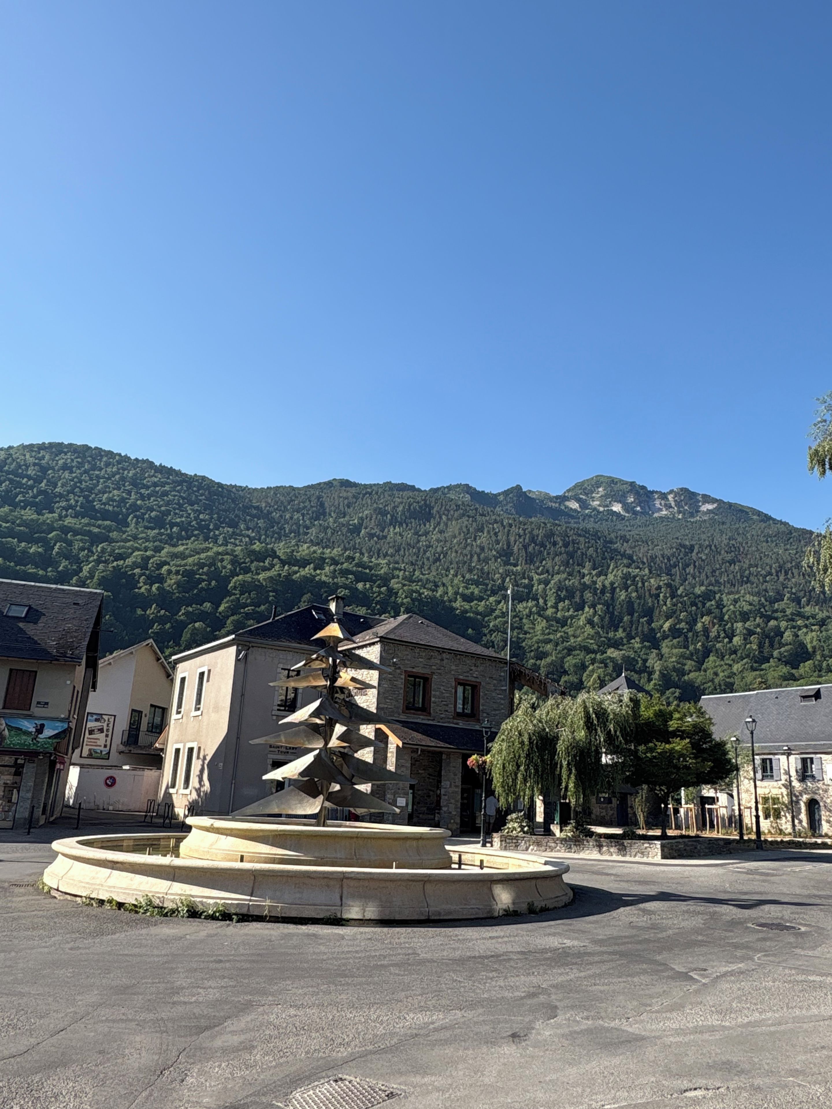 Una plaza de pueblo con una fuente central, rodeada de edificios de piedra y con montañas verdes al fondo.