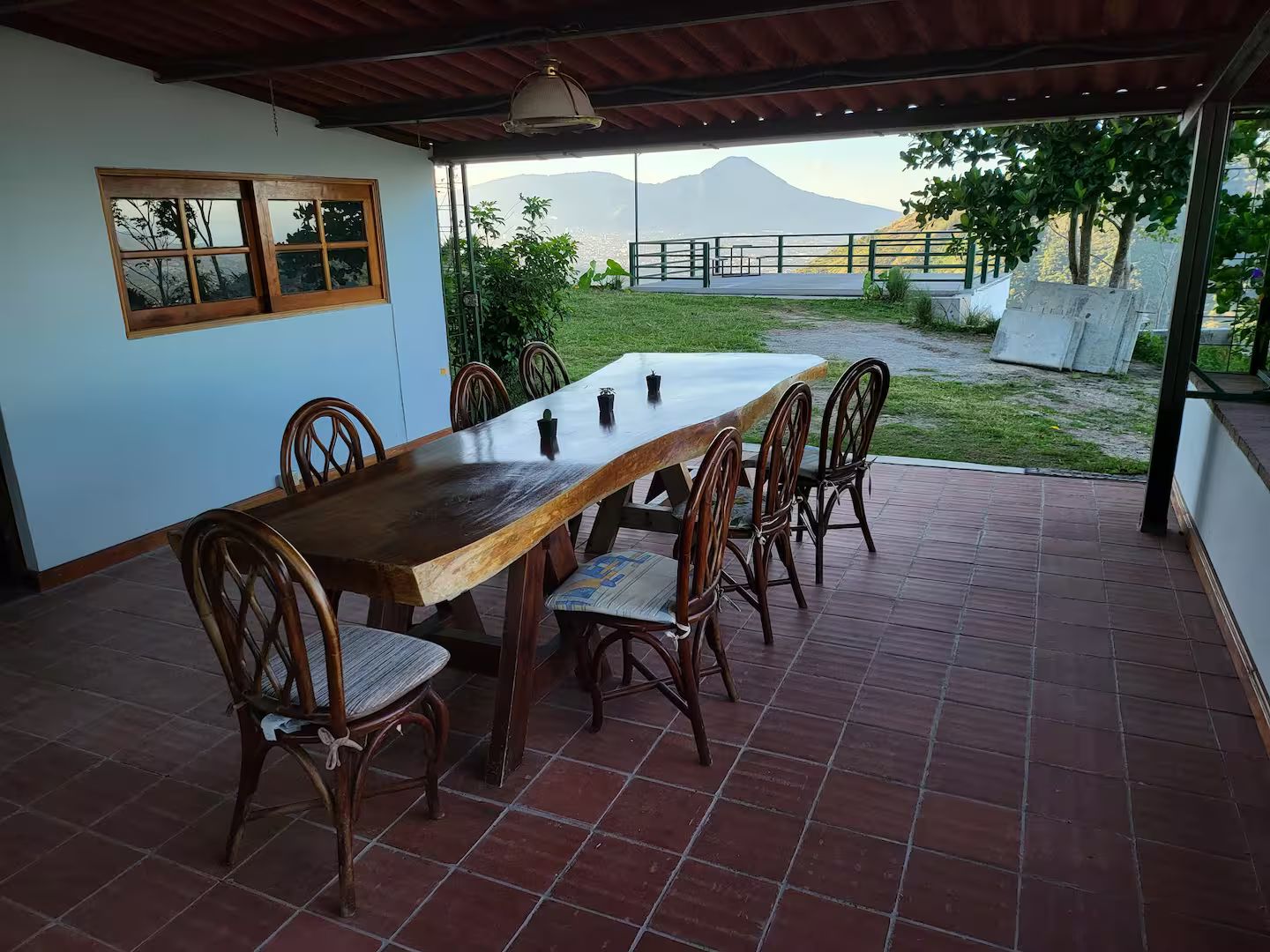 A covered outdoor dining area with a long wooden table and chairs, a view of a mountain, and a patio with a tiled floor.