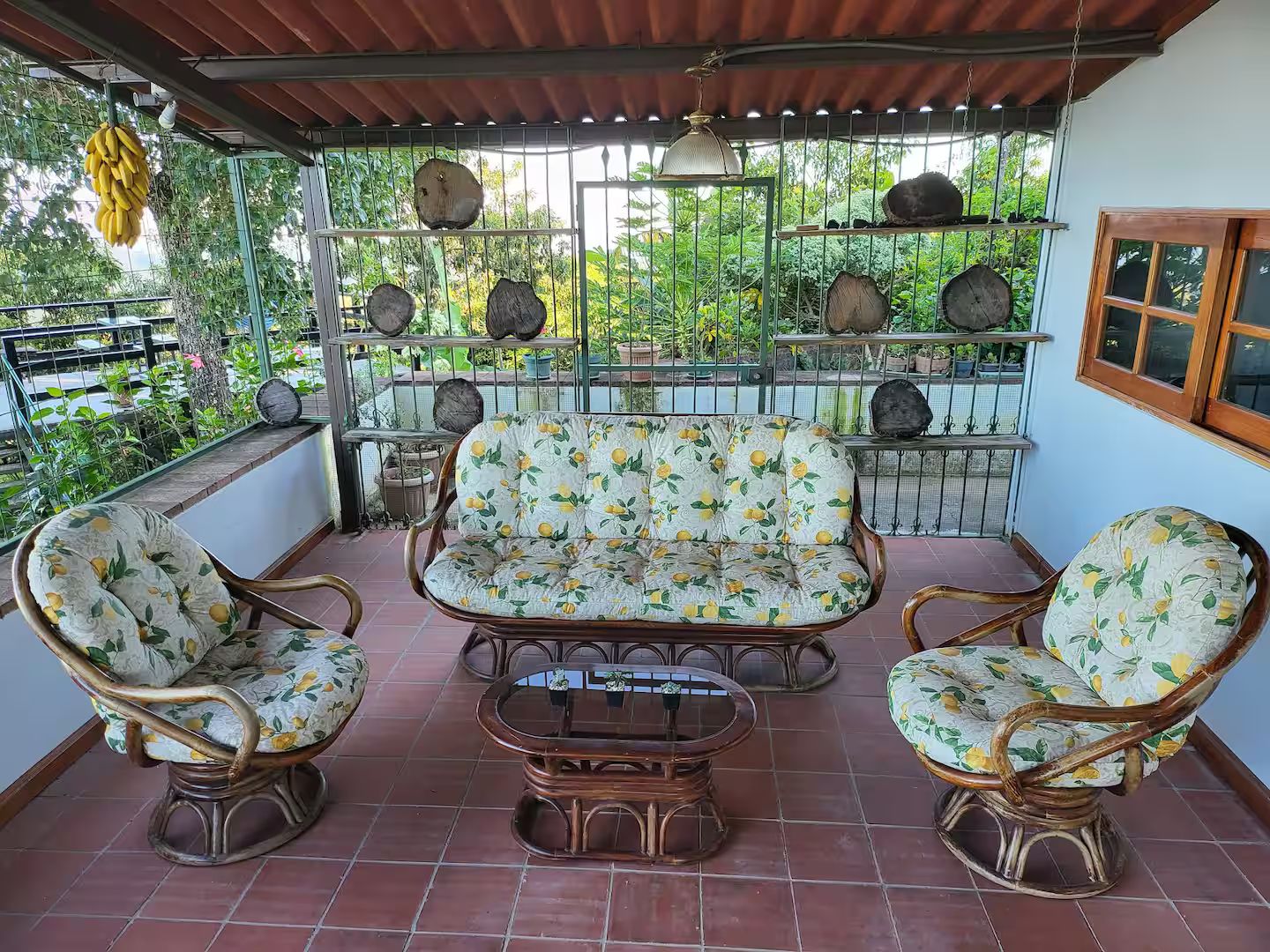 Ein überdachter Patio-Bereich mit Korbmöbeln, einem Glastisch und Blick auf Grün hinter einem Metallzaun.
