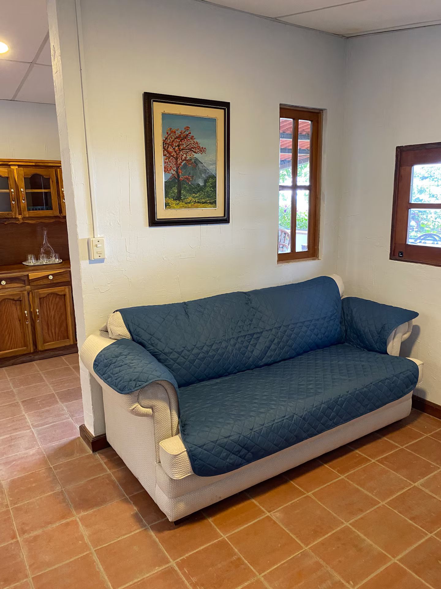 A room with a blue sofa, a framed painting, a window, and a kitchen area.