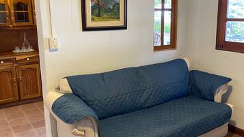 A room with a blue sofa, a framed painting, a window, and a kitchen area.