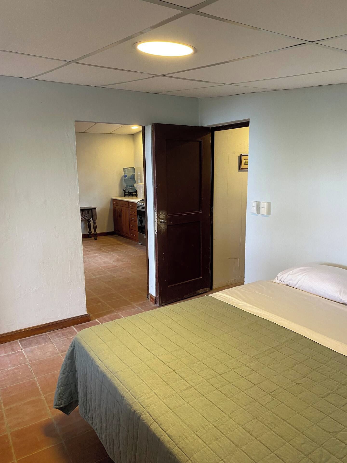 Habitación de hotel con cama, puerta, dispensador de agua y kitchenette.