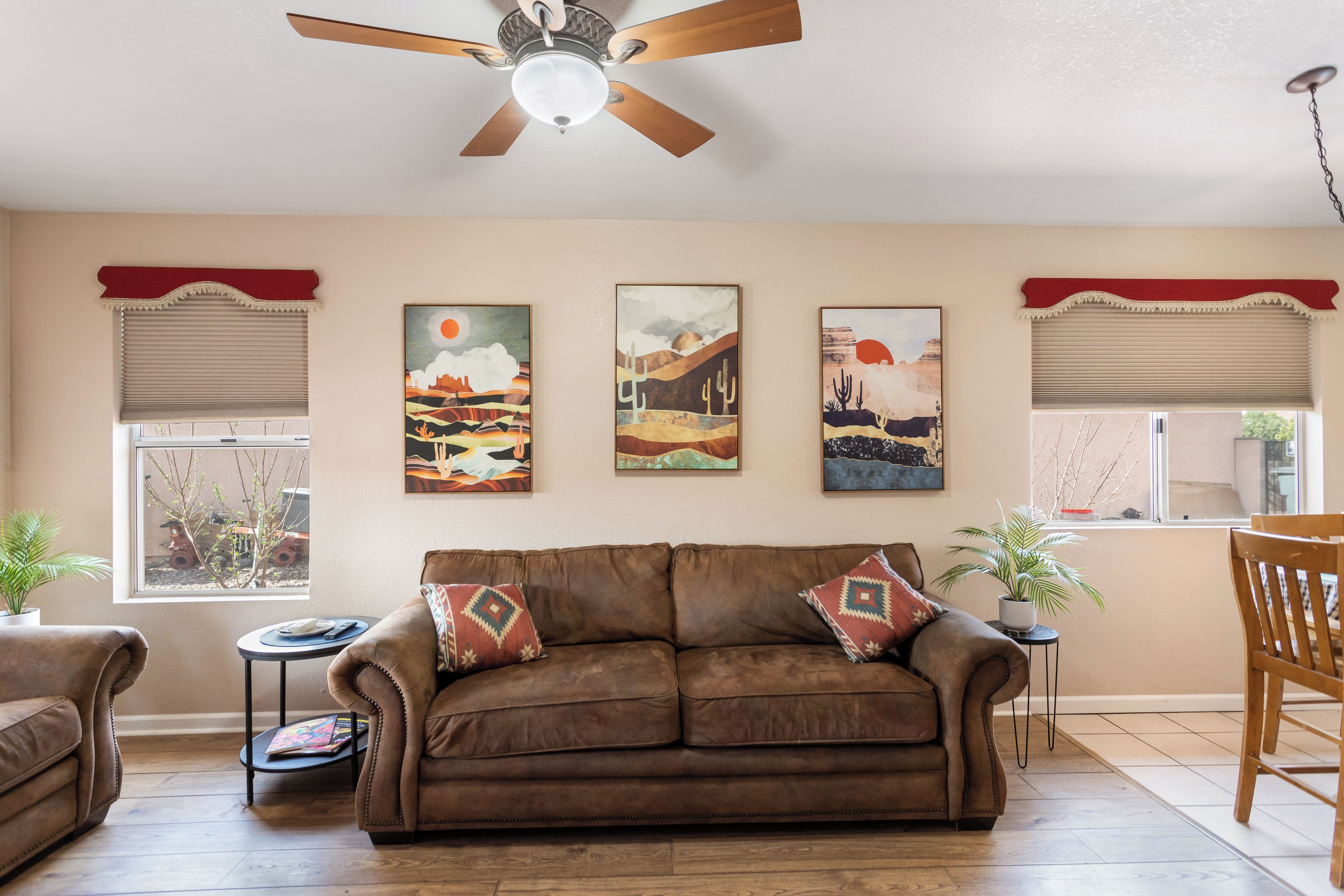 Un salon avec un canapé en cuir marron, un ventilateur de plafond et trois tableaux représentant des paysages accrochés au mur.