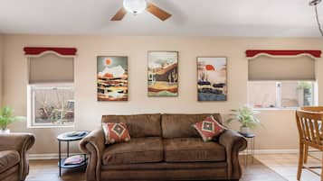 Un salon avec un canapé en cuir marron, un ventilateur de plafond et trois tableaux représentant des paysages accrochés au mur.