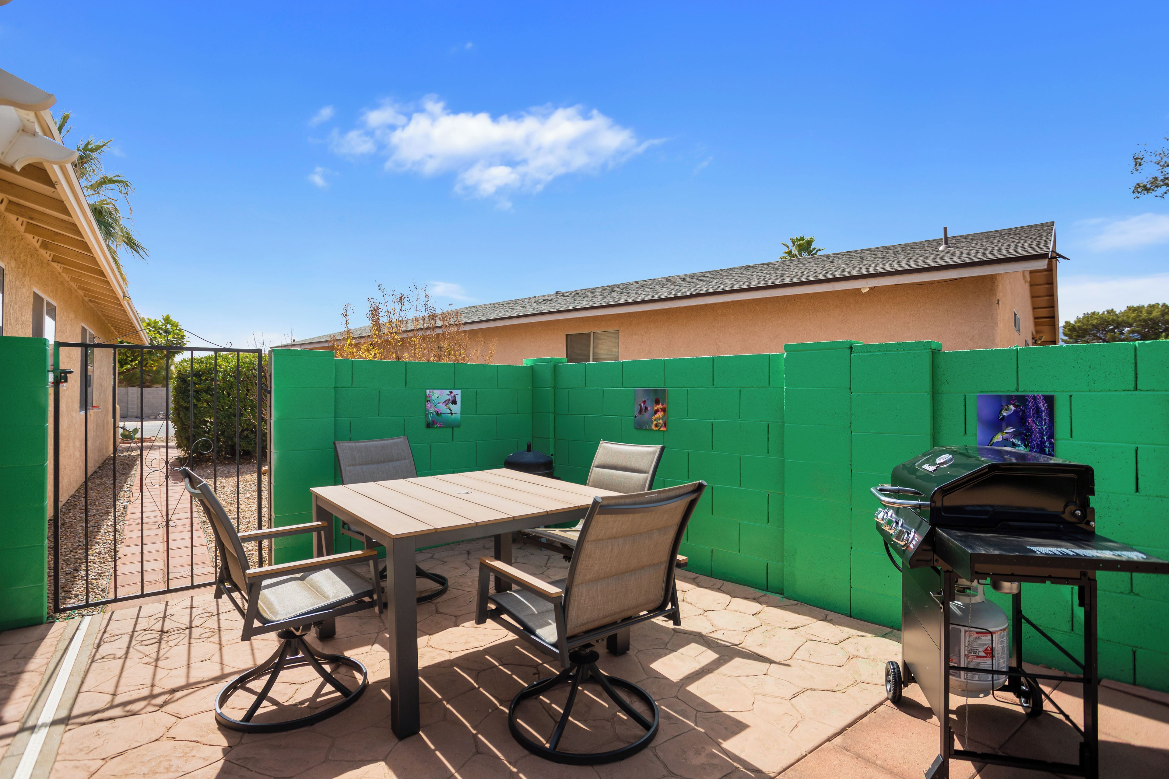 Patio esterno con tavolo e sedie, un barbecue e una recinzione verde.