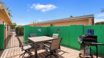 Patio esterno con tavolo e sedie, un barbecue e una recinzione verde.