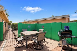 Patio esterno con tavolo e sedie, un barbecue e una recinzione verde.