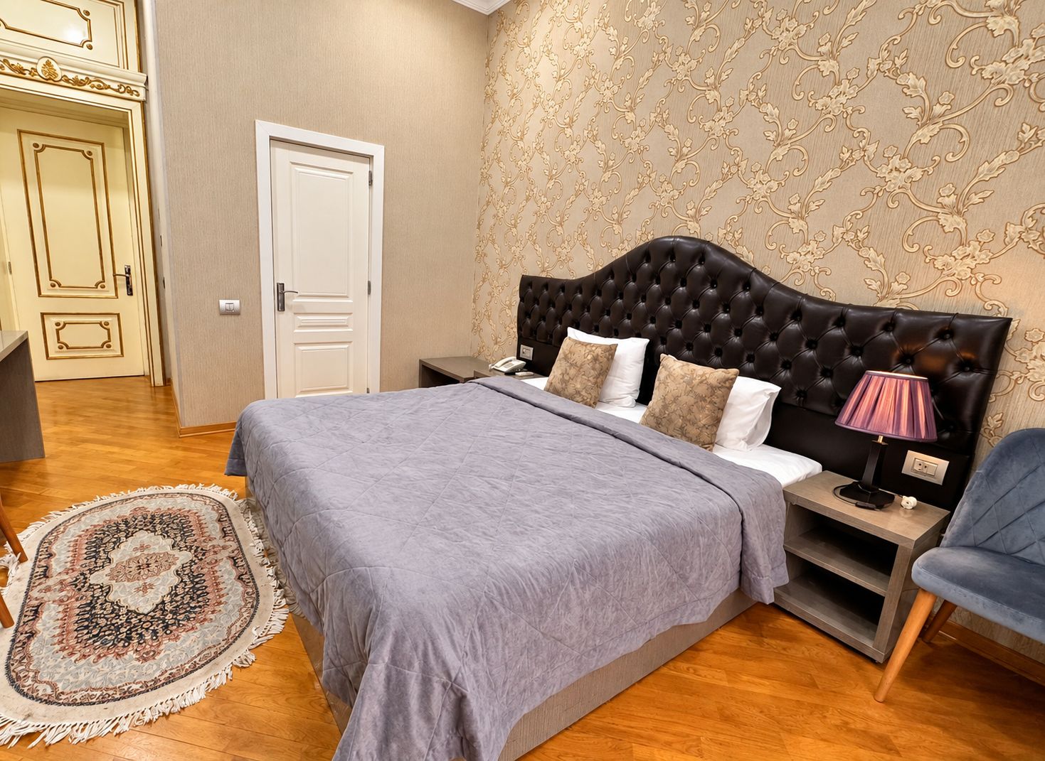 Une chambre à coucher avec une tête de lit capitonnée, un lit avec une literie grise, une lampe de chevet, un tapis à motifs et une porte ornée de moulures décoratives.