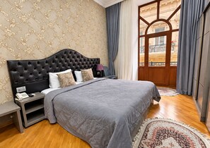 A bedroom with a tufted headboard, a bed with grey bedding, a bedside table with a lamp, and a view of a balcony through a wooden door. - Willdom Hotel (Baku)