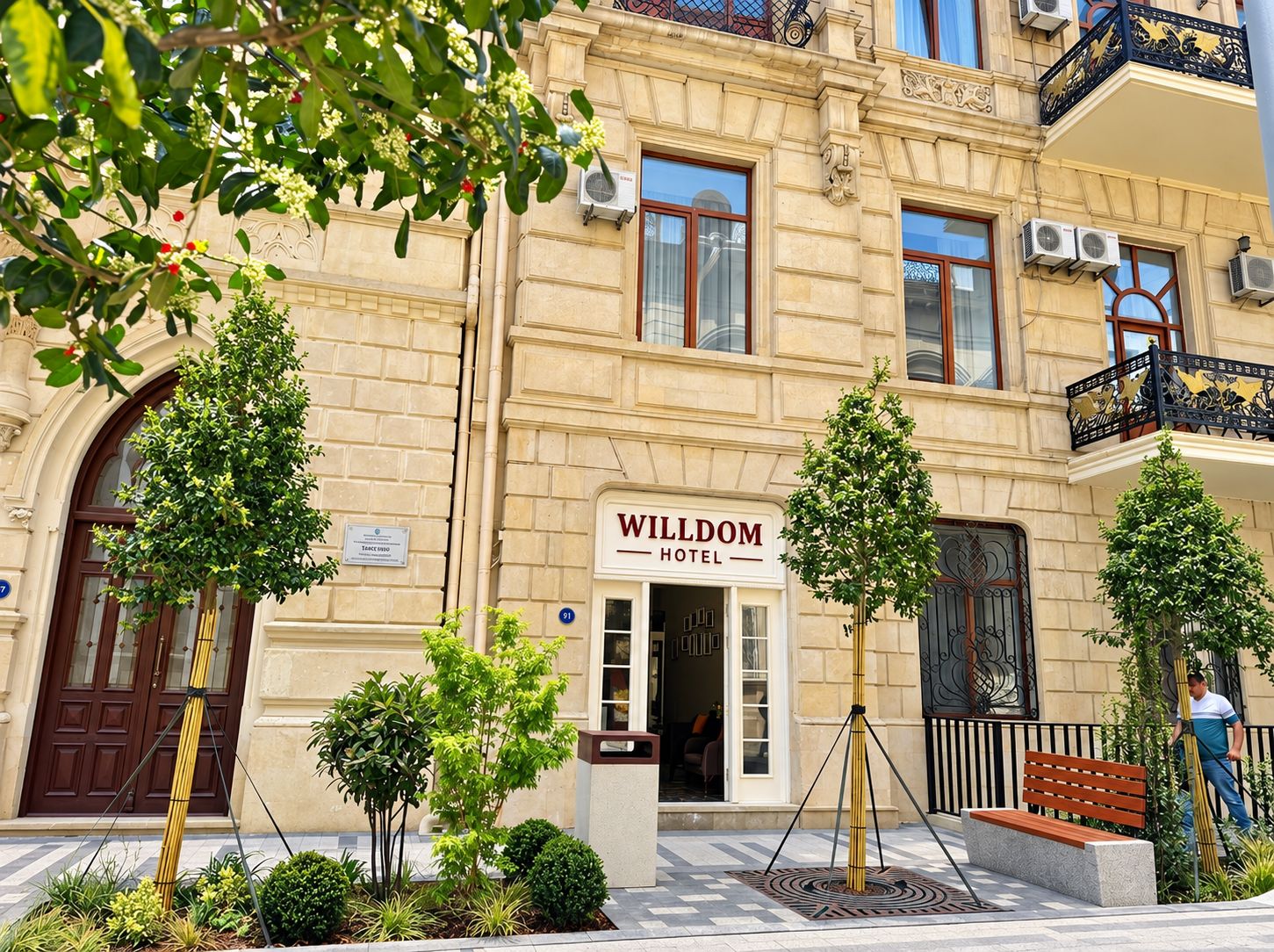 L’entrée d’un hôtel, agrémentée d’une enseigne indiquant « HÔTEL WILDOM », est entourée d’un aménagement paysager soigné et présente un style architectural traditionnel.