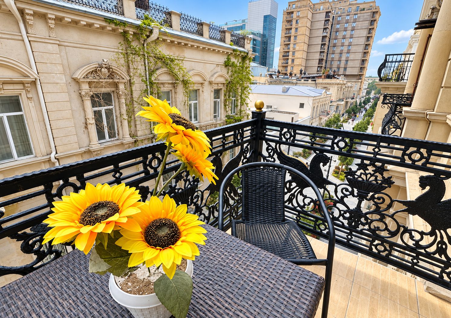 Un balcon orné d’une balustrade en fer forgé, une table en osier et un pot de fleurs contenant des tournesols.