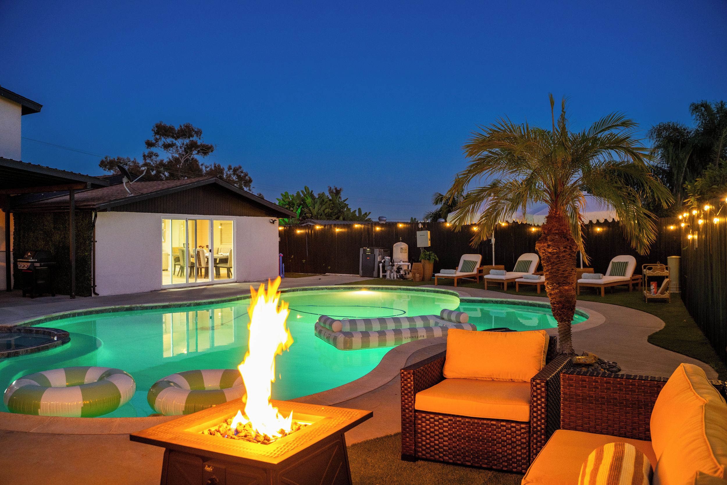A backyard with a swimming pool, a fire pit, lounge chairs, and a palm tree.