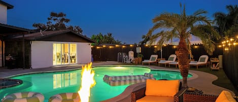 A backyard with a swimming pool, a fire pit, lounge chairs, and a palm tree.