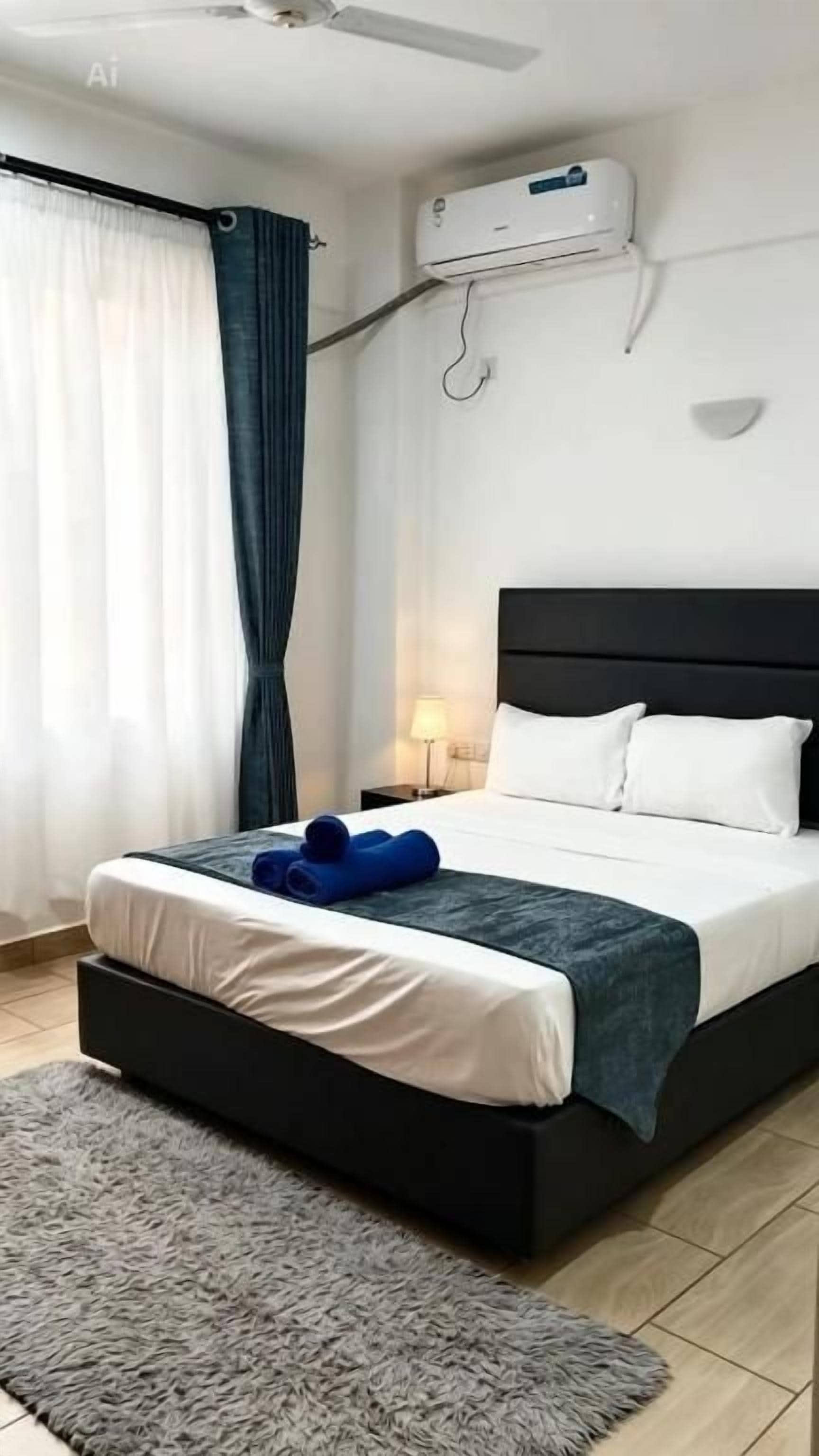 A hotel room with a bed, a nightstand, an air conditioning unit, and a window with curtains.