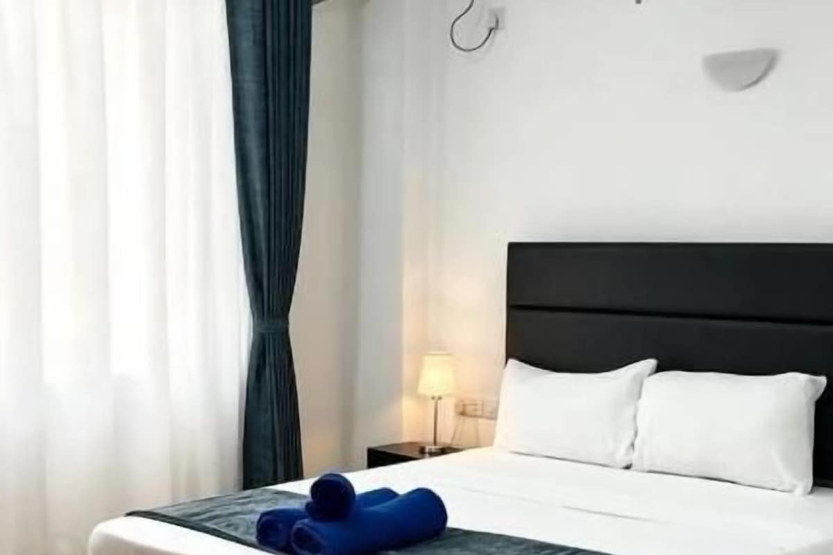 A hotel room with a bed, a nightstand, an air conditioning unit, and a window with curtains.