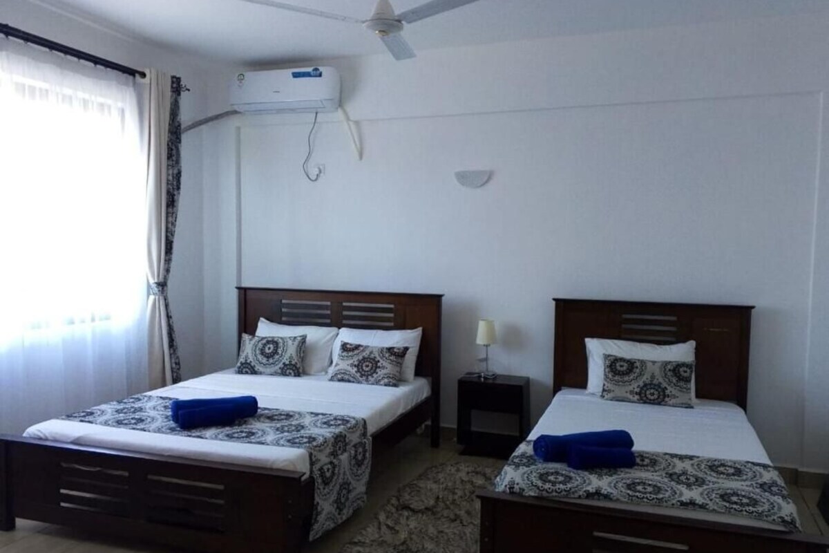 A hotel room with two beds, a ceiling fan, an air conditioning unit, and a small bedside table with a lamp.