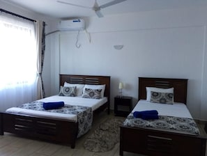 A hotel room with two beds, a ceiling fan, an air conditioning unit, and a small bedside table with a lamp.