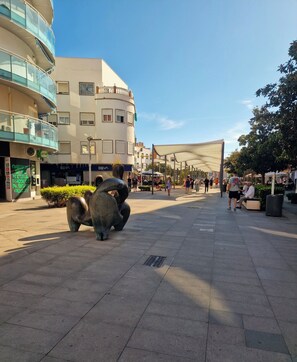 Una plaça pública amb una escultura gran, una zona de seients coberta i edificis amb botigues.