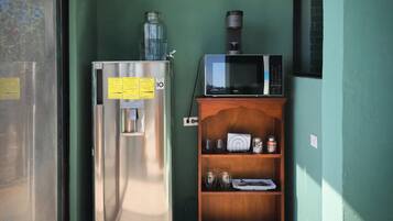 A compact kitchenette with a stainless steel refrigerator, a microwave, and a wooden shelf with various items.