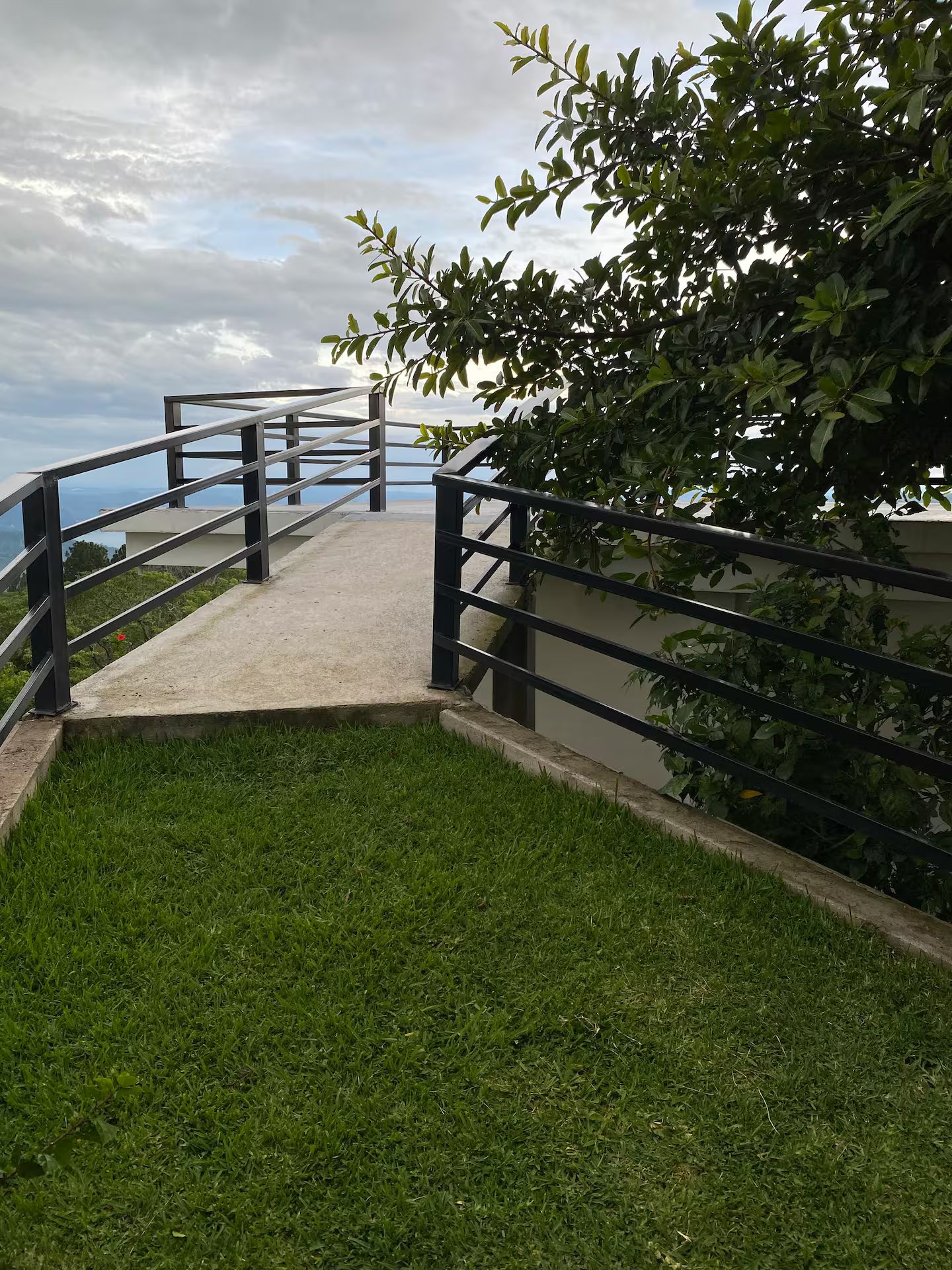 A concrete pathway with metal railings leading to a grassy area, surrounded by foliage.