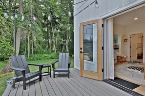 An outdoor deck with two Adirondack chairs, a small table, and a string of lights. - Fir Haven Farmhouse - Stylish 3BR Escape on Whidbey (Freeland)