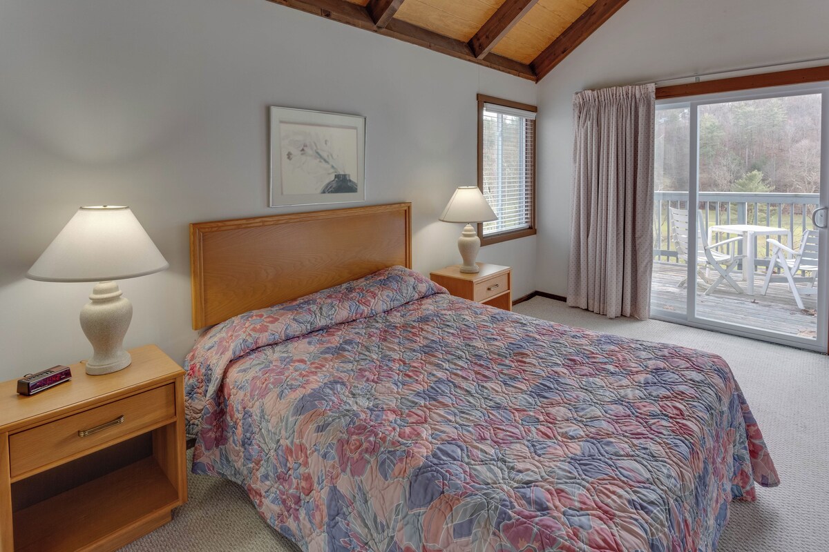 Un dormitorio con una cama, dos mesitas de noche, una mesita auxiliar, una ventana con persianas y vistas a una terraza exterior.