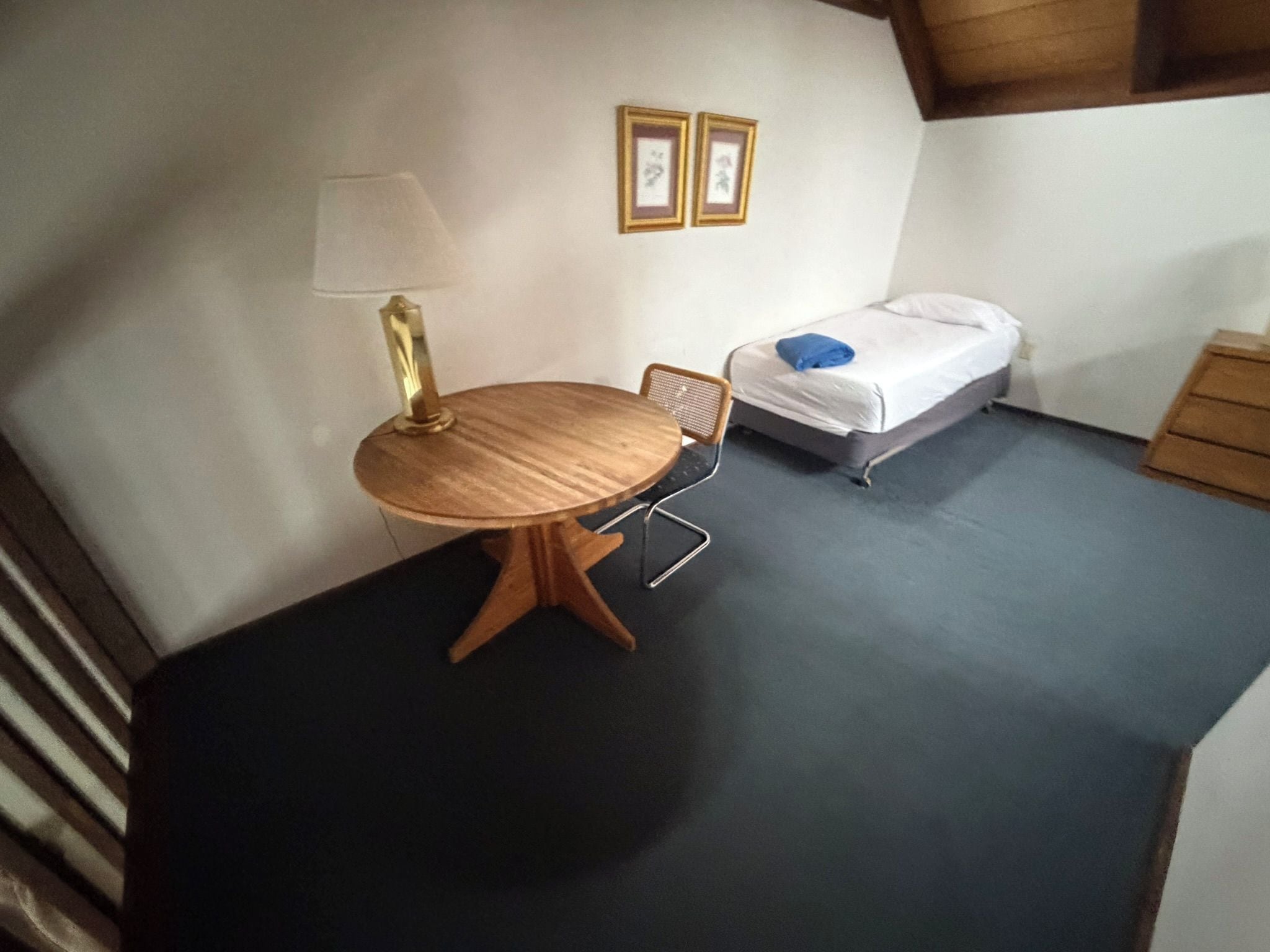 Une pièce de taille modeste, avec une table ronde en bois, une seule chaise, un lit avec un oreiller bleu et une petite table de chevet.